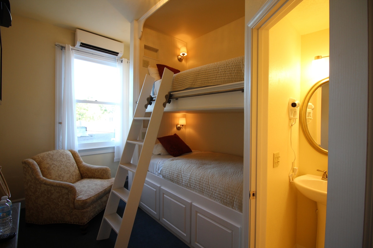 A cozy bunk room contains built-in twin bunk beds, complemented by a plush chair in one corner. A bathroom with a sink and mirror is adjacent to the sleeping area. Natural light enters through a window, and soft lighting is provided by wall sconces.