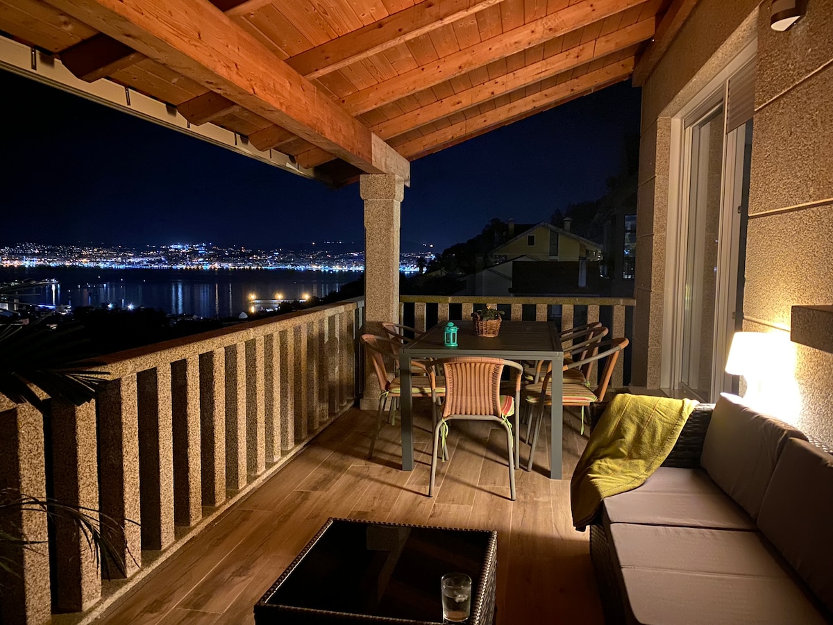 An outdoor terrace features comfortable seating and a dining table, all under a wooden roof. Soft lighting enhances the welcoming atmosphere, while stunning views of the city and bay of Vigo are visible in the background, creating an inviting space for relaxation.