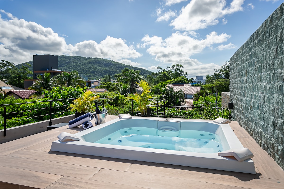 A rooftop spa is featured, surrounded by a wooden deck and lush greenery. The spa's clear water reflects the sunlight, with a backdrop of distant hills under a partly cloudy sky. Towels are neatly placed on the side, enhancing the inviting ambiance.