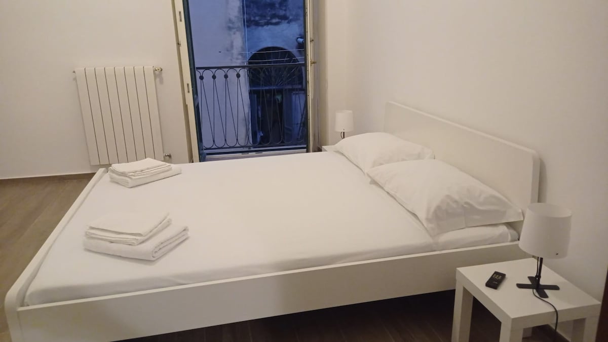 A bright sleeping area features a spacious white bed with neatly arranged bedding. A small nightstand with a lamp complements the minimalist design. Natural light filters in through an open door that leads to a balcony, enhancing the room's airy ambiance.