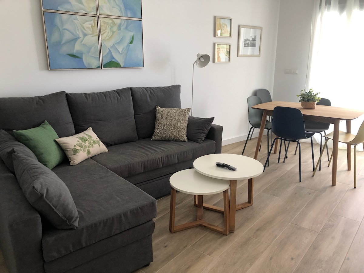A modern living area is showcased, featuring a spacious gray sectional sofa adorned with decorative pillows. A light-colored coffee table is positioned in front, complemented by a round dining table with various seating options. Natural light streams in through the nearby window.