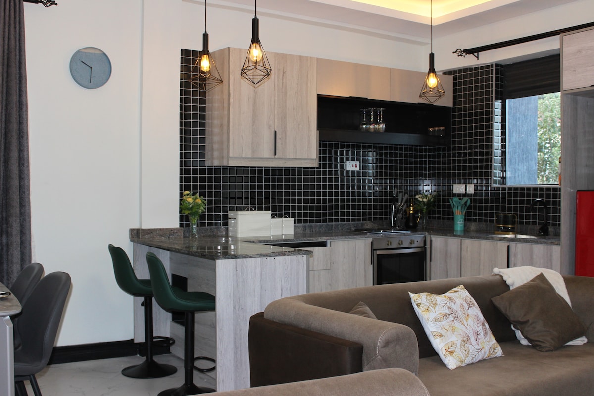 A modern kitchen area is showcased with sleek cabinets and a dark tiled backsplash. A well-equipped stove and refrigerator are visible alongside a round dining table with four chairs. Pendant lighting adds a subtle glow, enhancing the atmospheric appeal of the space.