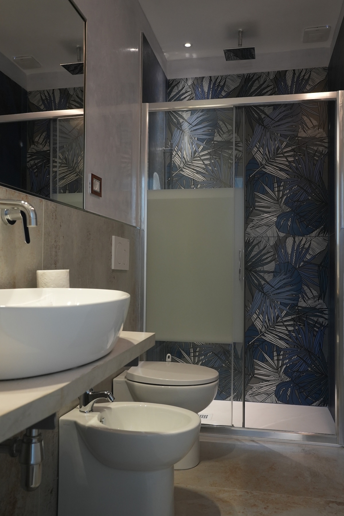 A modern bathroom featuring a circular sink mounted on a smooth countertop. A shower space is enclosed with glass panels, displaying a decorative blue and green leaf-patterned wall. A white toilet sits adjacent to the sink, with soft lighting providing a welcoming atmosphere.
