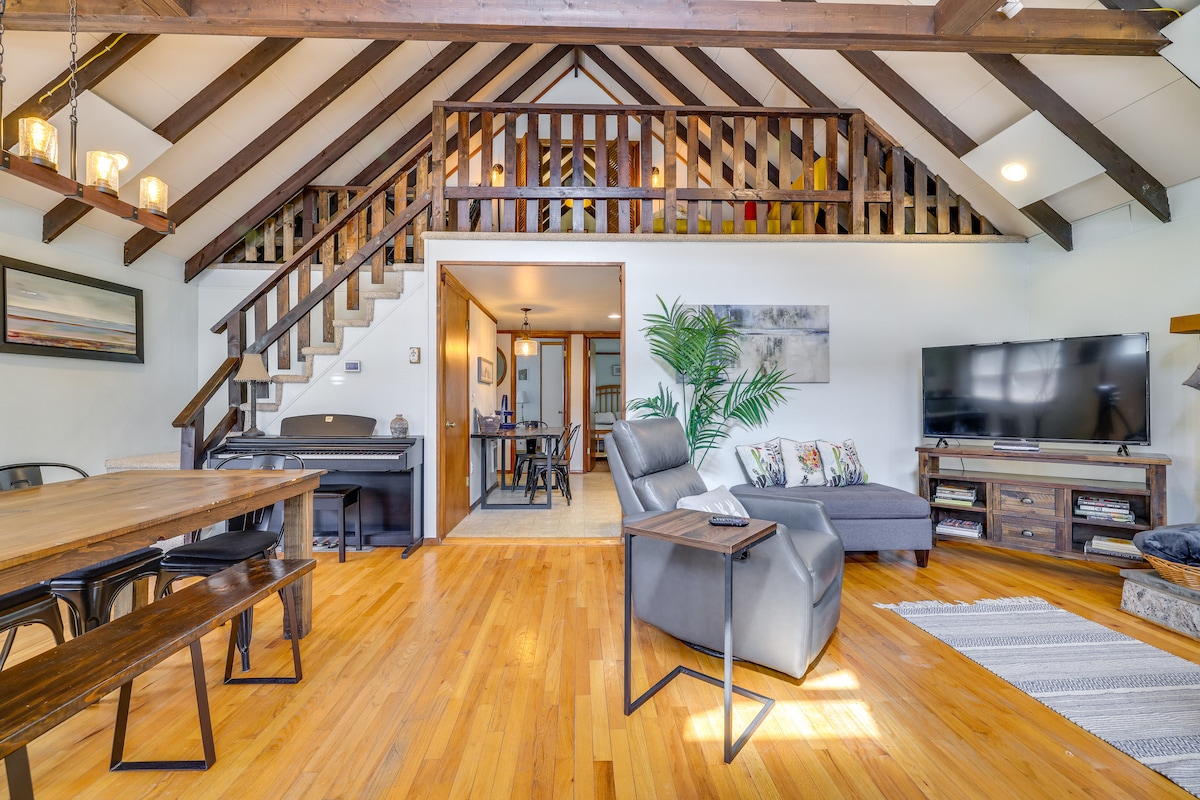 An open living space is highlighted by exposed wooden beams and a mix of cozy furniture. A stone fireplace and a large smart TV are visible, accompanied by a dining area featuring a long wooden table. Natural light illuminates the warm wooden floors.