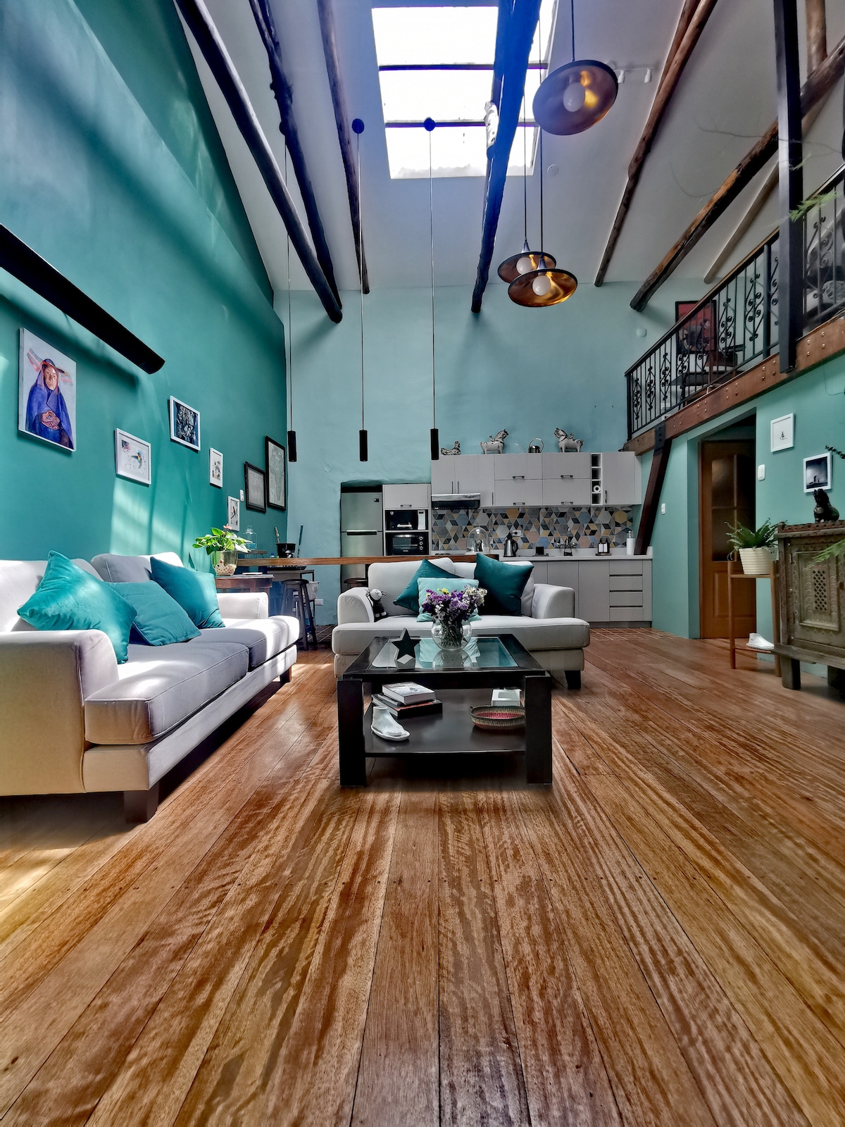 A spacious living area features high ceilings and large windows that allow natural light to fill the room. Comfortable couches with teal cushions are arranged around a glass coffee table. A mix of modern and decorative elements can be seen on the walls and shelves.