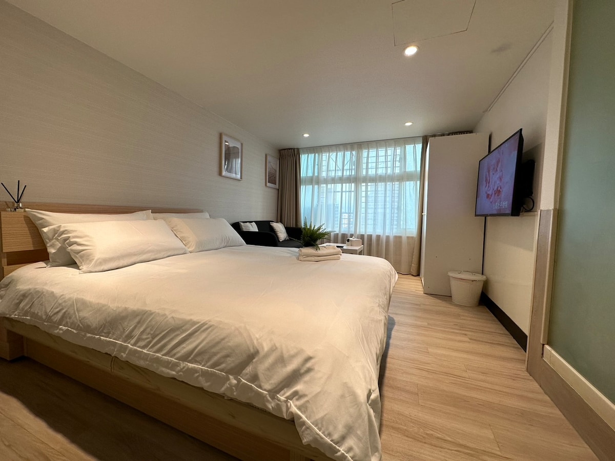 A spacious sleeping area features a large bed covered with crisp white linens. Soft natural light is let in through large windows, complemented by a cozy seating area with a dark couch. A flat-screen TV is mounted on the wall opposite the bed.