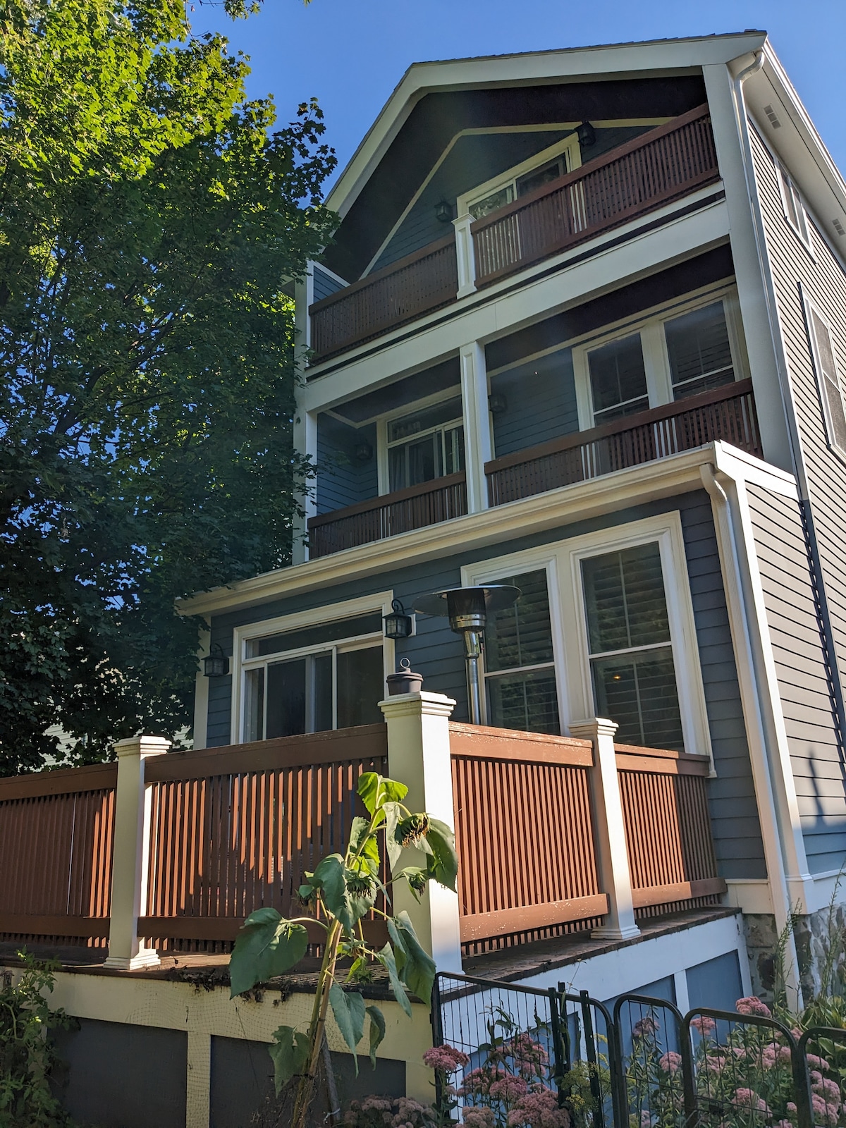 A three-story home with wooden siding and multiple balconies is surrounded by lush greenery. The front features a covered porch with a railing and lanterns, providing a welcoming entrance. Sunlight filters through the trees, creating a natural ambiance.