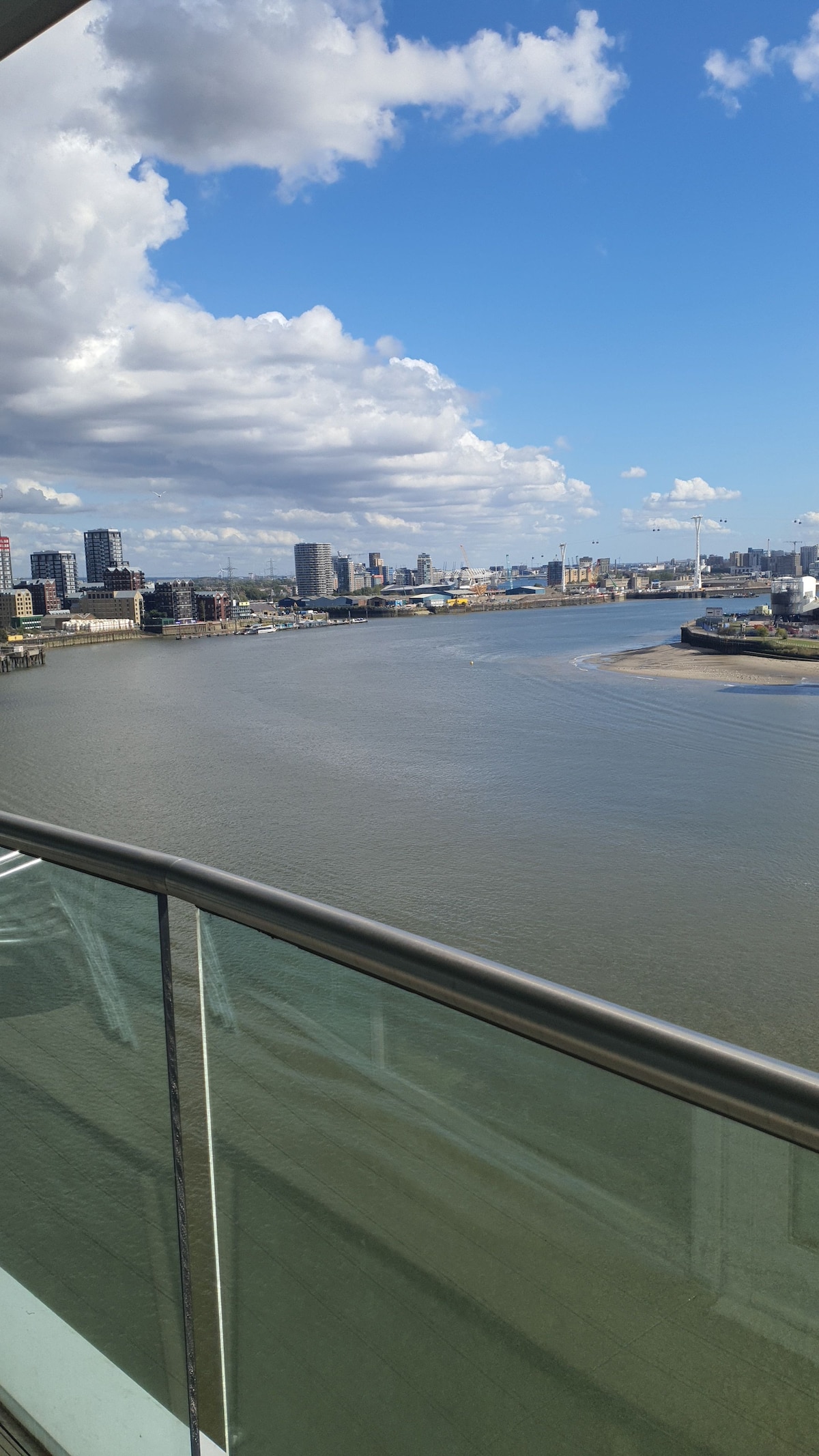 A panoramic view of the Thames River is captured from a balcony, showcasing the city skyline with various buildings and structures along the waterfront. Fluffy clouds drift across a bright blue sky, reflecting off the water's surface.