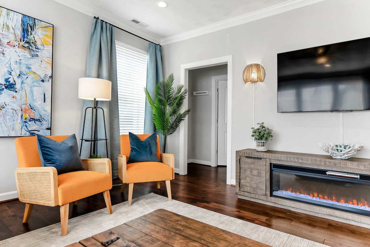 A bright living area is showcased with two vibrant orange chairs and a stylish floor lamp. A large flat-screen TV is mounted on the wall, and a decorative plant adds a touch of greenery. A modern electric fireplace offers a cozy focal point beneath the TV.