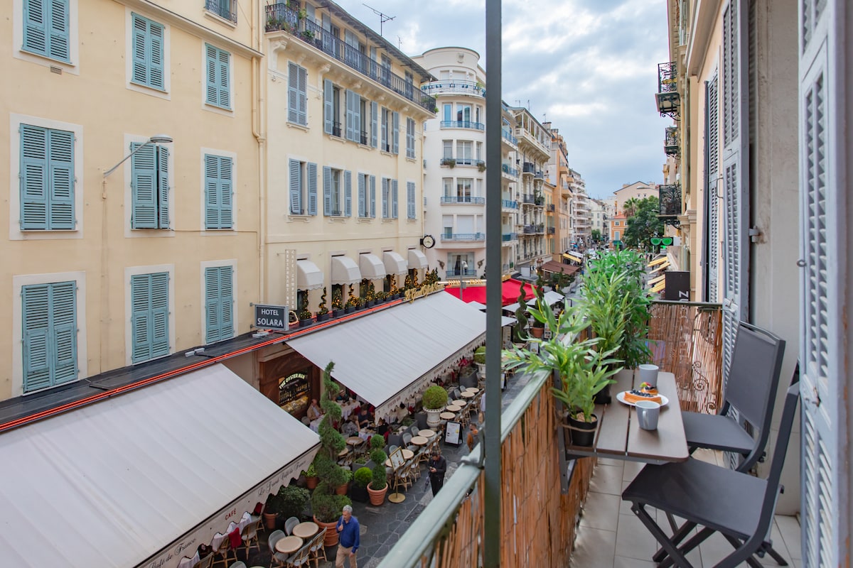 A balcony view overlooks a lively street lined with charming buildings. Tables and chairs are arranged on the outdoor patio below, with umbrellas providing shade. Lush greenery adorns the balcony railing, complementing the inviting atmosphere of the surrounding restaurants.