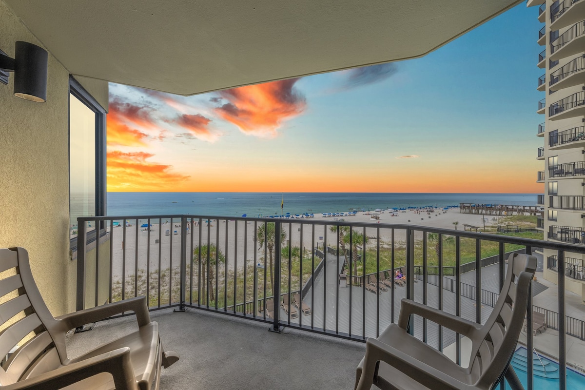 A spacious balcony offers panoramic views of the beach and ocean, where vibrant sunset colors blend in the sky. Two comfortable chairs are positioned for relaxing, with the sandy beach and umbrellas in the distance, creating a serene coastal atmosphere.