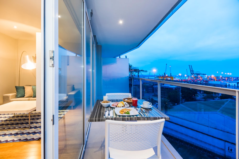 A private balcony offers a table set for breakfast, featuring a variety of dishes, with a view of the port illuminated by evening lights. A cozy seating area is visible through the glass doors, enhancing the inviting ambiance of the space.