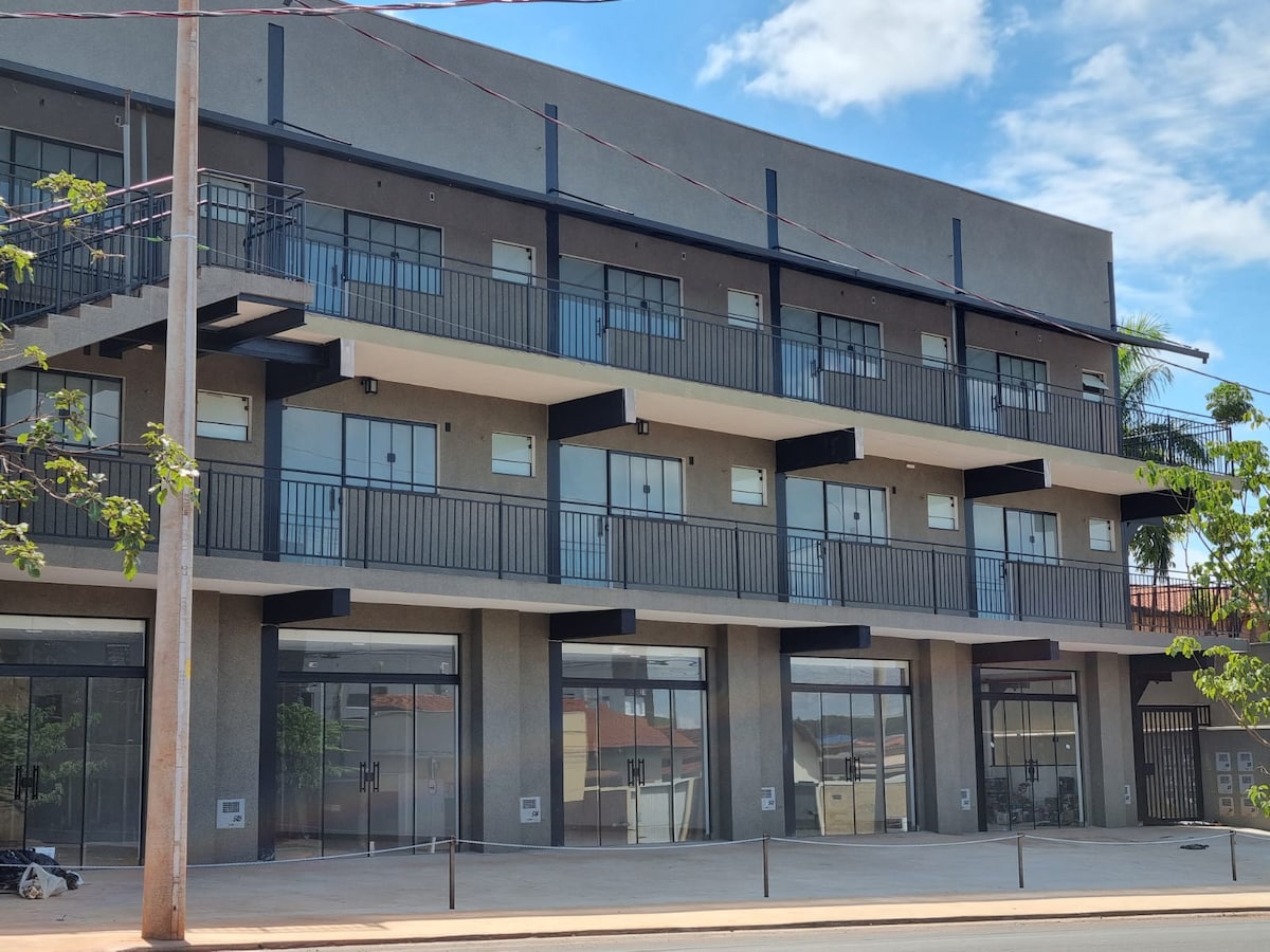 A modern building with multiple levels features a uniform façade and balconies on the upper floor. Large glass entryways are visible on the ground level, allowing natural light to illuminate the entry space. The surroundings include greenery and a clear blue sky.
