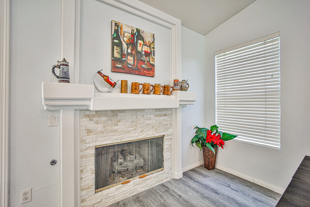A welcoming living area is centered around a modern fireplace with a stone facade. Decorative items, including wooden mugs and a framed artwork, are displayed on the mantel. A window with vertical blinds allows natural light to illuminate the space, complemented by a potted plant.