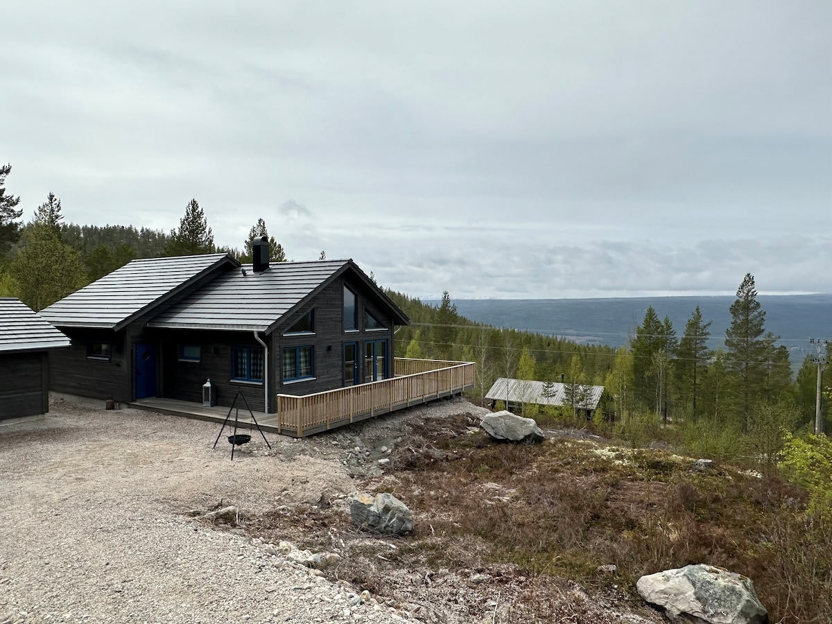 A modern wood cabin situated on a hillside, surrounded by trees and natural vegetation. The cabin features a sloped roof and a deck that offers a view of distant mountains and valleys, enhancing the serene landscape. Gravel paths lead to the structure, adding to the rustic charm.