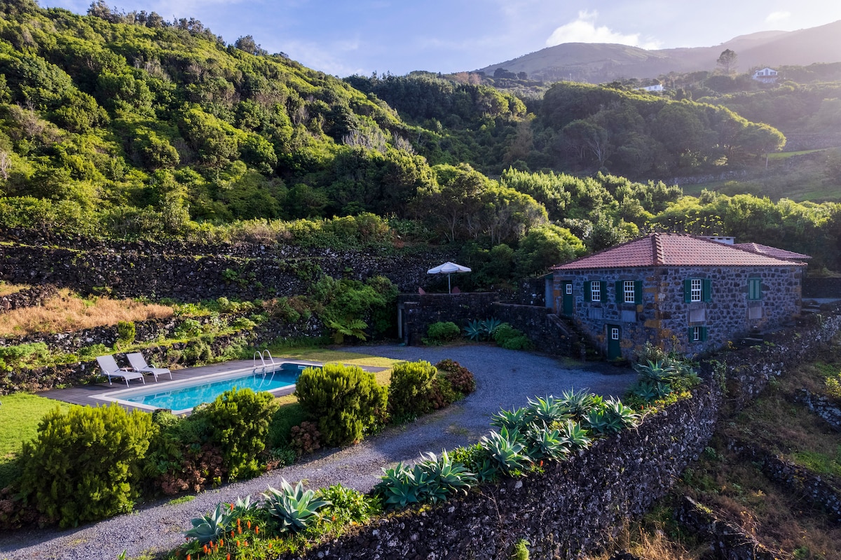 A view of a stone house surrounded by lush greenery, featuring a vibrant outdoor pool with sun loungers. The property is framed by terraced landscaping, and a shaded umbrella offers a resting spot. Rolling hills can be seen in the background under a clear sky.