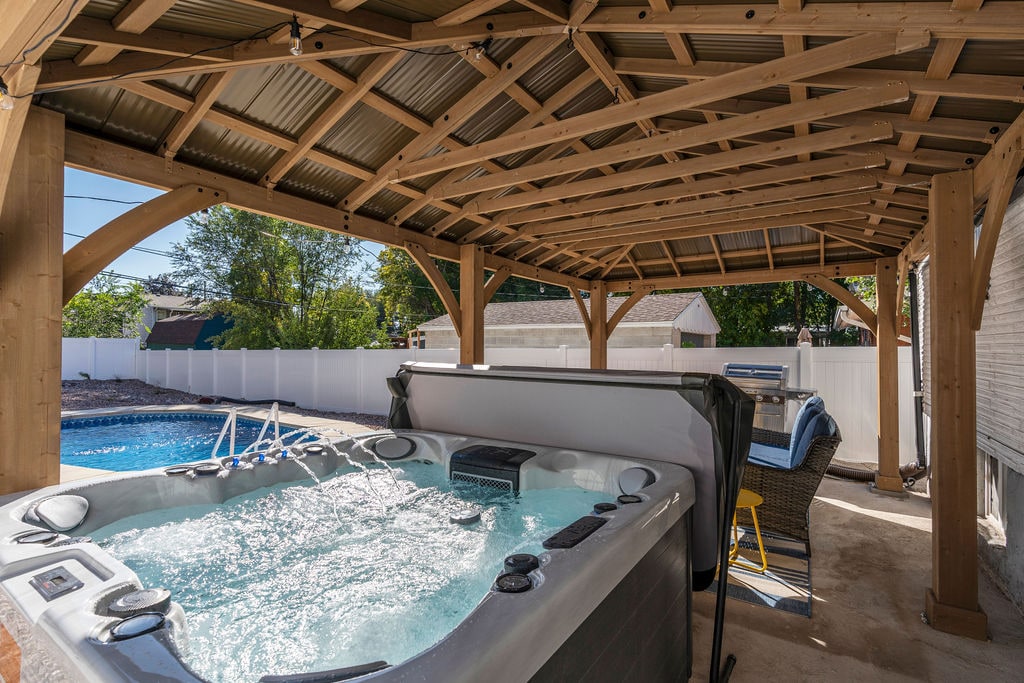A covered outdoor space features a spacious hot tub, with bubbling water inviting relaxation. Nearby, comfortable lounge chairs are positioned under a wooden structure, providing shade. A clear view of the swimming pool can be seen in the background, surrounded by a white fence.