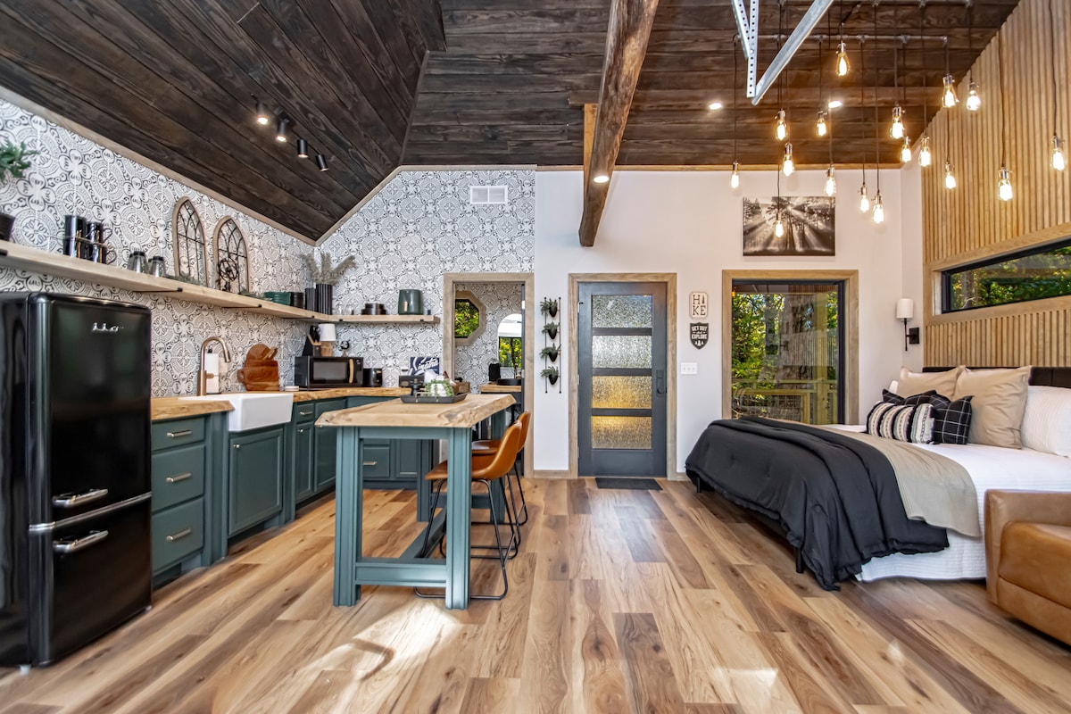 An elegant interior features wooden beams and stylish pendant lighting. A spacious kitchen area includes a dark refrigerator, complemented by a dining table and modern chairs. A cozy sleeping area is furnished with a large bed, with natural light flowing through large windows, enhancing the inviting space.