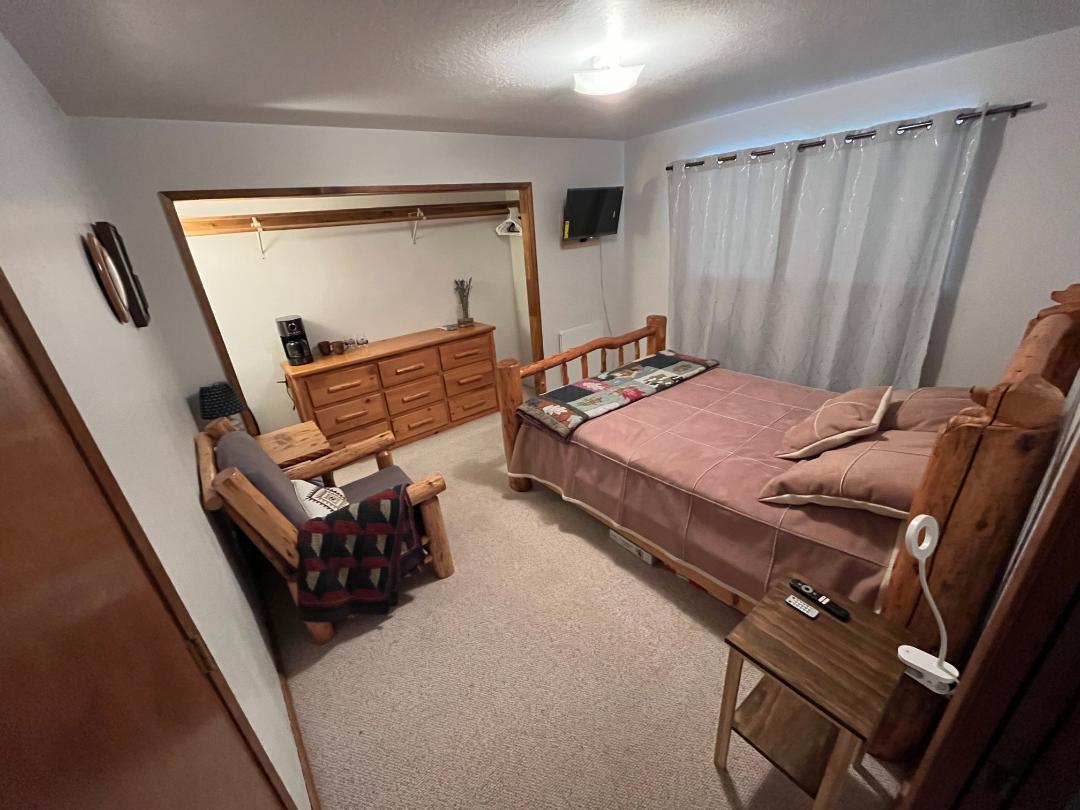 A cozy bedroom features a spacious log bed with a soft blanket and pillows. A natural wood armchair sits nearby, and a dresser with multiple drawers is positioned against the wall. Light curtains allow for soft lighting, while a small TV is mounted for convenience.