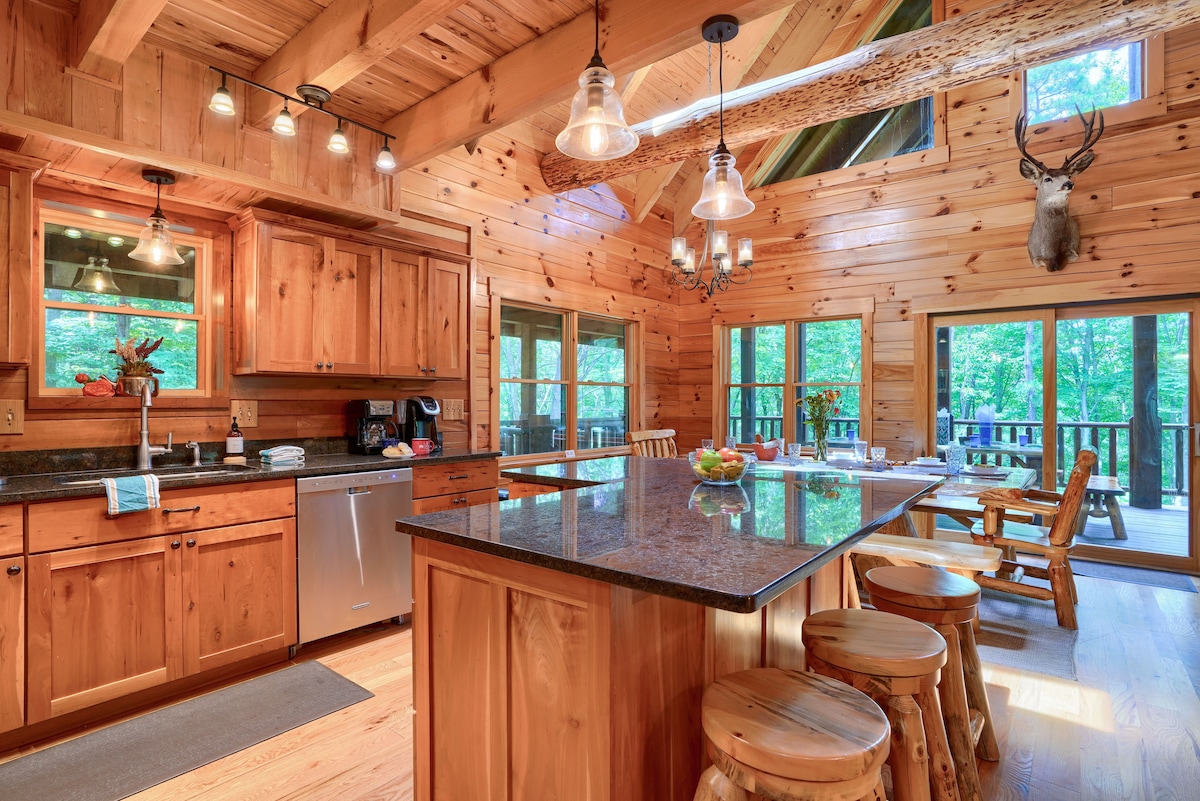 The kitchen area is crafted with natural wood cabinetry and features stainless steel appliances. A spacious island, topped with dark granite, provides ample space for meal preparation. Windows frame views of the surrounding woods, offering natural light to the inviting atmosphere.