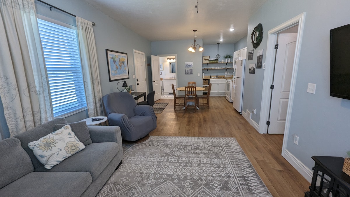 A spacious living area features a comfortable gray sofa and a soft area rug, creating a cozy ambiance. Natural light enters through the window, illuminating the adjacent dining area with a wooden dining table and chairs. The kitchen is visible in the background, showing modern appliances.