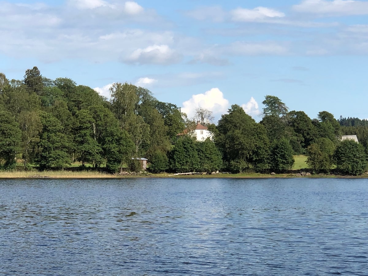The image displays a scenic view of a serene lake bordered by lush greenery. A charming white building is positioned amidst trees in the background, reflecting the natural beauty of the surrounding area. Bright blue skies adorned with soft clouds enhance the tranquil atmosphere.
