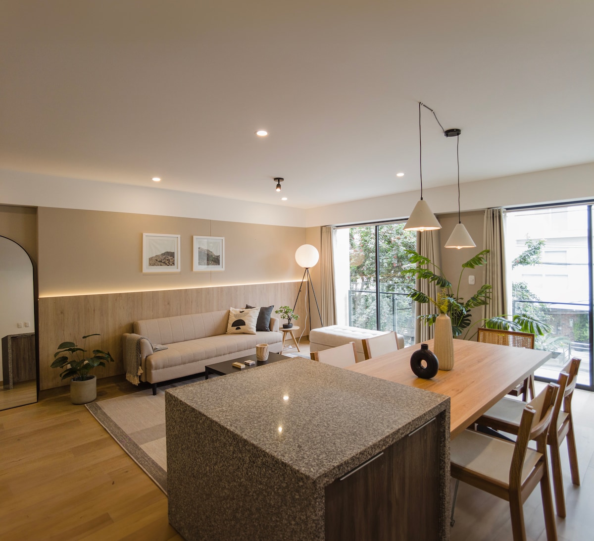 An open plan living and dining area features large windows allowing natural light to fill the space. A cozy sofa is positioned across from a wooden dining table surrounded by chairs. A decorative floor lamp and plants add a touch of warmth to the modern setting.