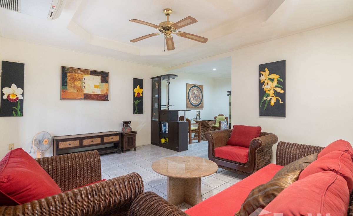 A spacious lounge area is featured with comfortable seating, including two red cushioned sofas and a round coffee table. Wall art depicting flowers adds color to the neutral walls, while a ceiling fan provides airflow. An adjoining space is visible, showcasing the dining area.