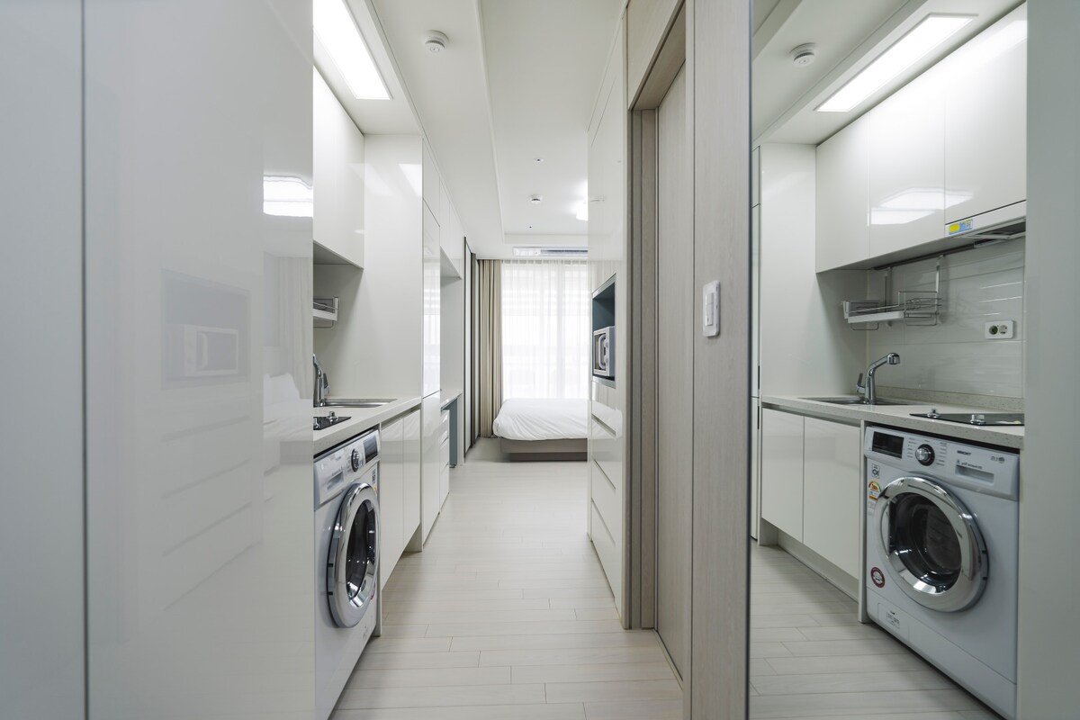 A modern kitchen area is visible, featuring sleek cabinetry and essential appliances, including an induction stove and washing machine. Soft light filters through a large window, illuminating the pathway to a spacious bedroom in the background, creating an airy and functional layout.