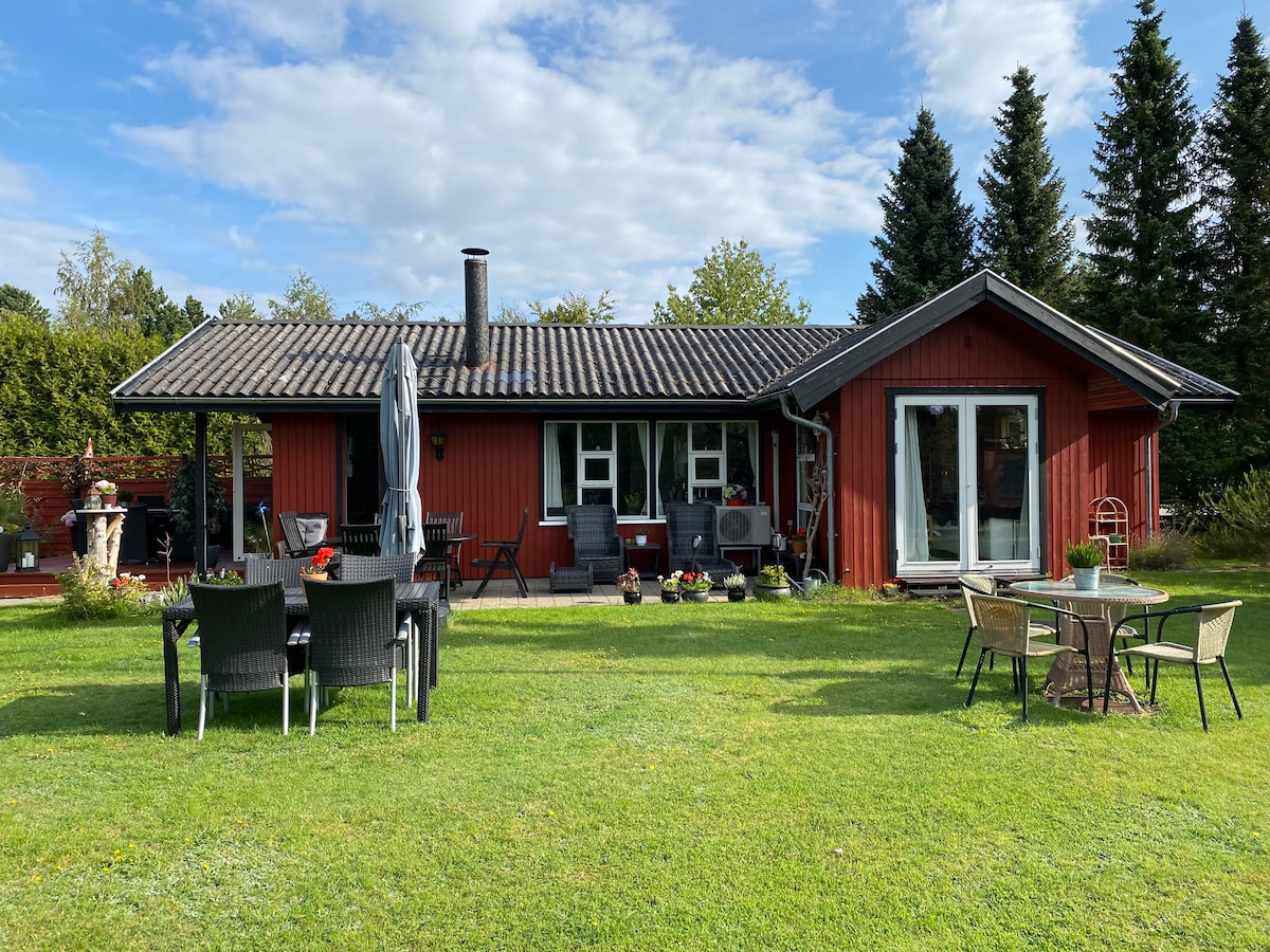 A red wooden house is set amidst well-kept greenery, featuring large windows and a covered patio. Outdoor seating areas include a table with chairs and a separate lounging space. Surrounding trees offer shade and privacy, contributing to a peaceful outdoor environment.