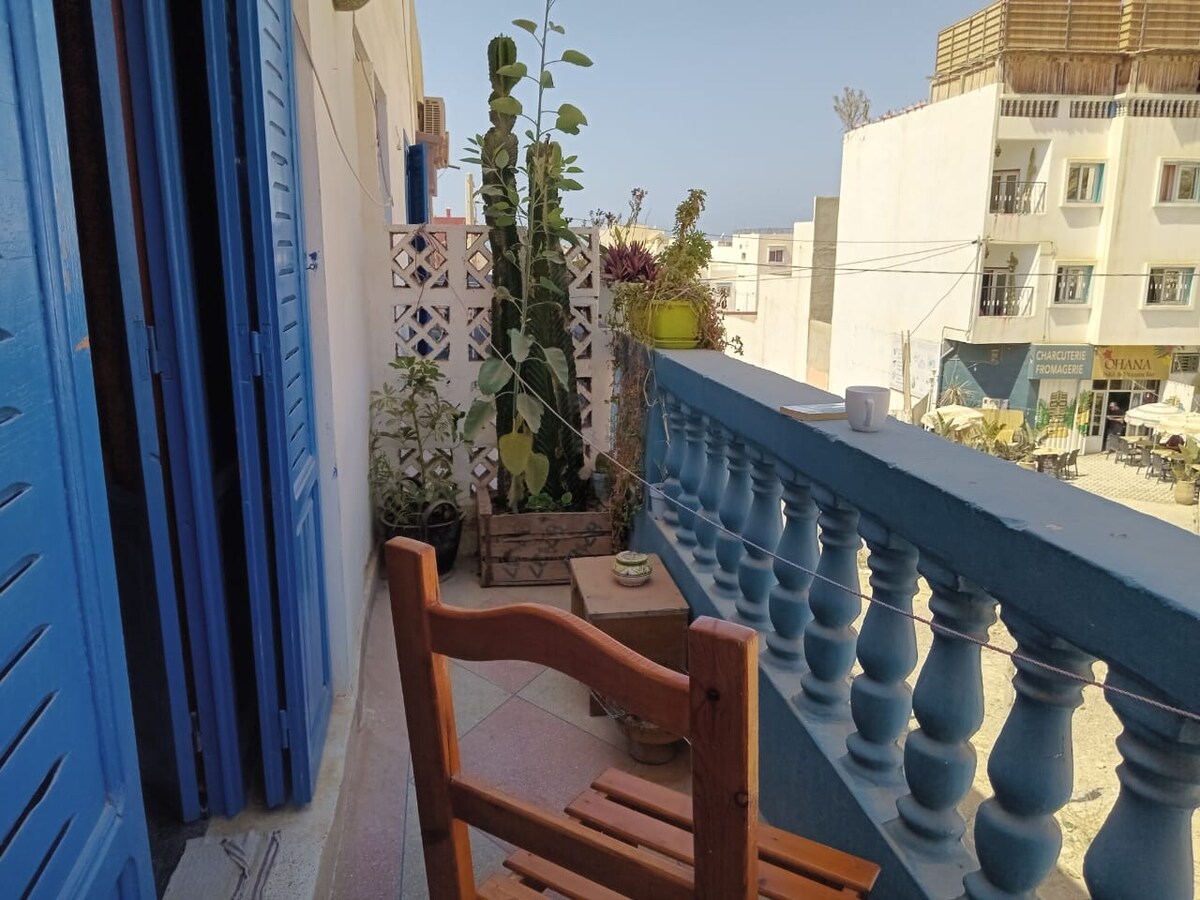 A sunny balcony features blue railings and overlooks a view with neighboring buildings. A wooden chair and small table are positioned on one side, complemented by various potted plants, creating a relaxing outdoor space for enjoying morning coffee.