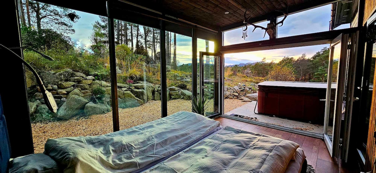 A spacious sleeping area features large glass walls that open to a natural outdoor setting, revealing landscaped stones and greenery. Soft bedding is neatly arranged on the bed, providing a serene view of the surrounding landscape and the setting sun.