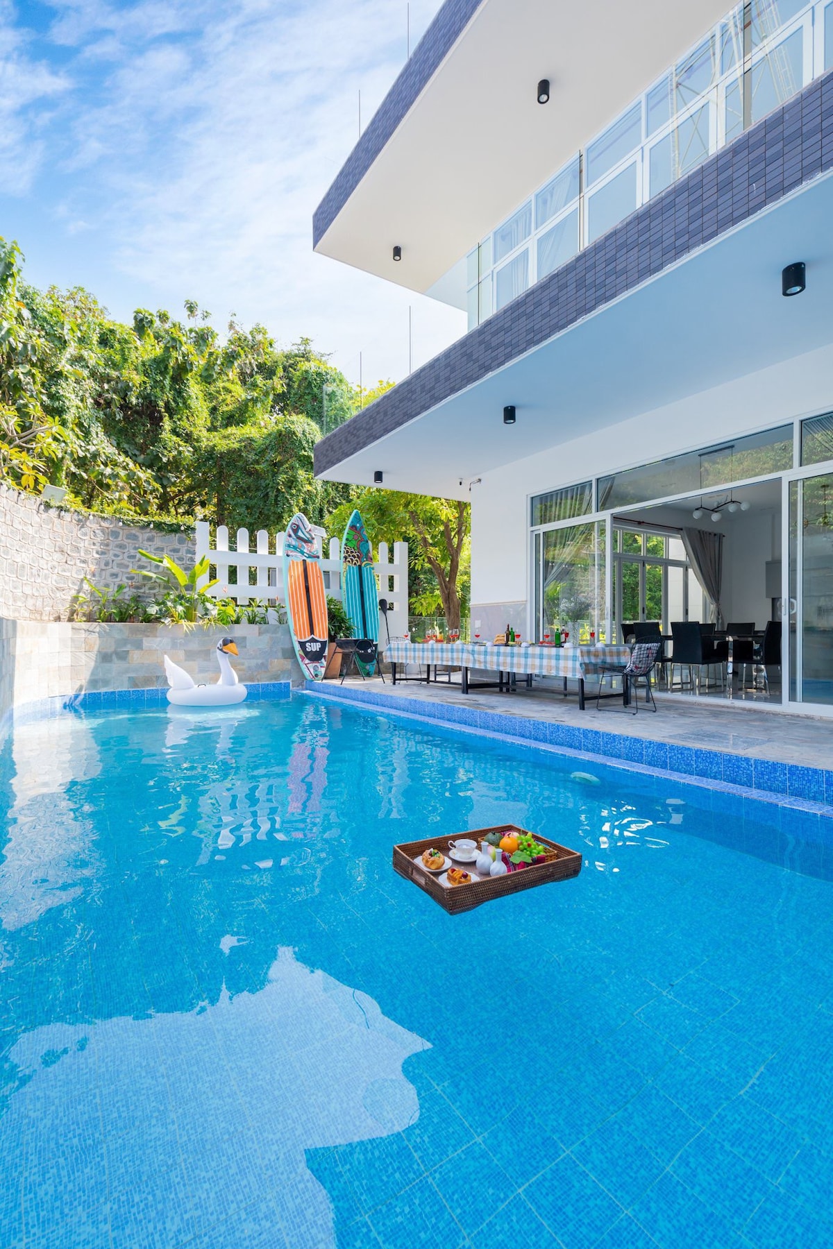 A tranquil swimming pool reflects the blue sky, framed by a modern villa. A wooden tray with refreshments floats in the pool. Surfboards stand against the wall, while outdoor seating is arranged nearby, inviting relaxation in a serene environment.