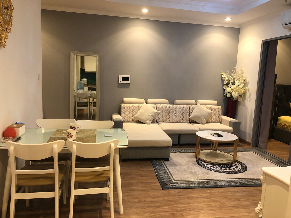 A spacious living area is featured, showcasing a light-colored sectional sofa paired with a round coffee table. A dining table with four chairs is positioned nearby. Soft lighting illuminates the room, which is decorated with elegant floral arrangements. A mirror reflects the space.