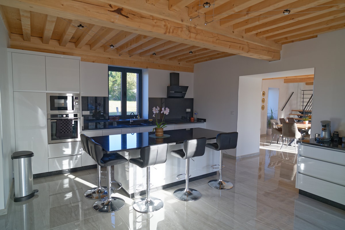 A modern kitchen is equipped with sleek white cabinetry and black countertops. Bar stools with black seats are arranged around the central island. Large windows allow natural light to fill the space, highlighting the wooden ceiling beams above. An orchid adds a touch of greenery.