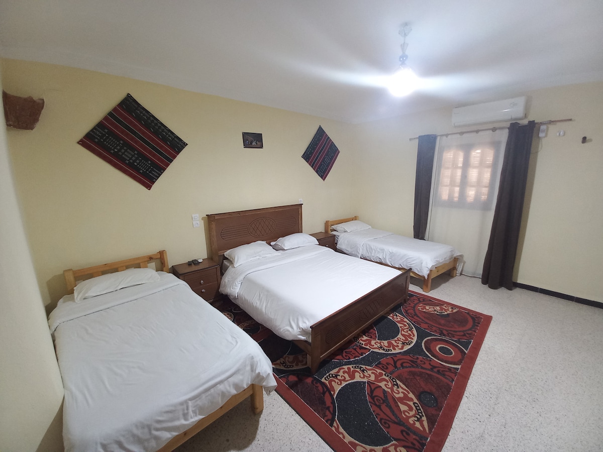 A spacious hotel room features a large double bed and a single bed, accompanied by an additional single bed. Colorful traditional wall hangings are displayed above the beds. A patterned area rug covers the floor, and natural light enters through a window dressed with dark curtains.