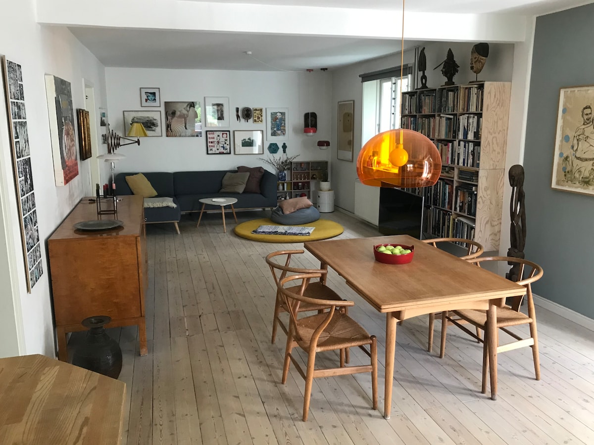 A spacious living area is presented, featuring a comfortable sofa and various art pieces on the walls. A dining table with four chairs occupies the center, complemented by a bowl of fruit. Shelving units and an orange pendant light add character to the space.