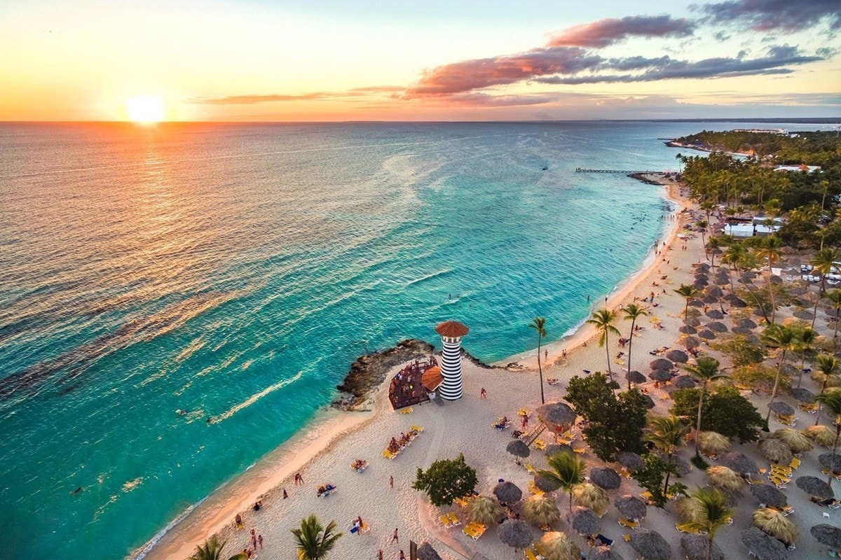 An aerial view of a beach at sunset presents an expansive coastline. Gentle waves lap against the shore, where umbrellas and lounge chairs are arranged. A horizon of palm trees and a brightly colored lighthouse add to the tranquil scene, with the sky displaying a gradient of warm colors.