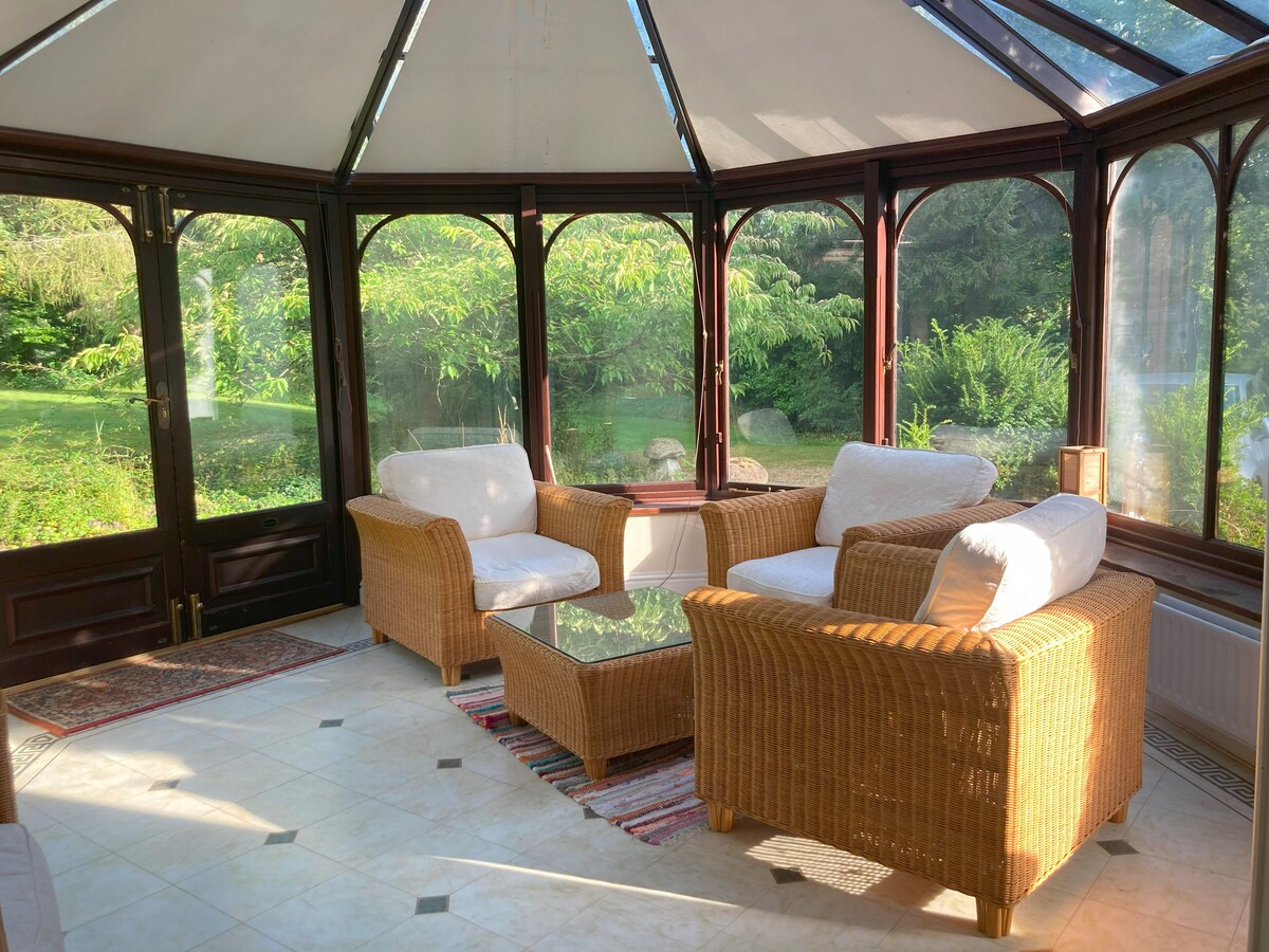 A bright conservatory features wicker seating with white cushions arranged around a glass coffee table. Large windows offer views of a lush garden, allowing natural light to illuminate the space. A colorful rug adds texture to the tiled floor.