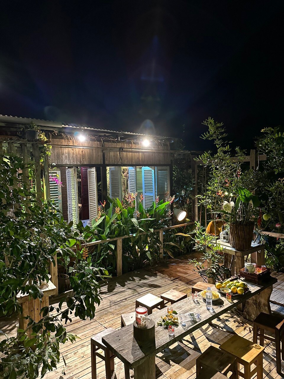 An outdoor seating area is illuminated by warm lights, featuring a rustic wooden table surrounded by chairs. Lush greenery frames the space, enhancing the natural ambiance. The structure showcases wooden walls and colorful shutters, creating a cozy and inviting atmosphere for gatherings.