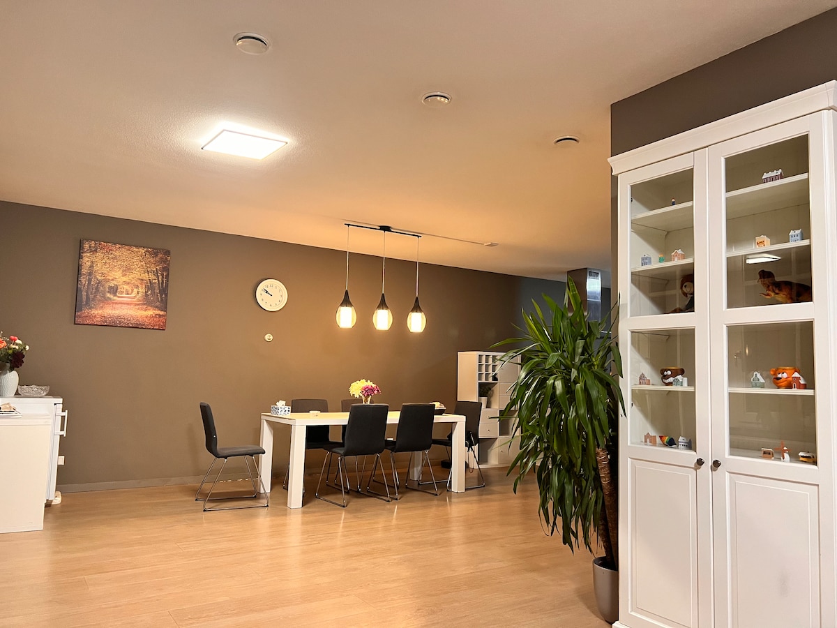 A spacious dining area is featured, illuminated by pendant lights above a central table surrounded by black chairs. A decorative clock and a landscape artwork adorn the wall. A glass-front cabinet displays decorative items, while a potted plant adds a touch of greenery.