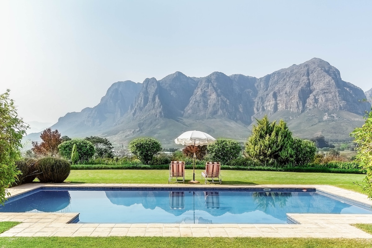 An outdoor pool is surrounded by a well-maintained lawn and lush greenery, offering views of the distant mountains. Two lounge chairs are positioned beneath a white umbrella, providing a shaded area for relaxation. The serene landscape emphasizes the connection with nature.