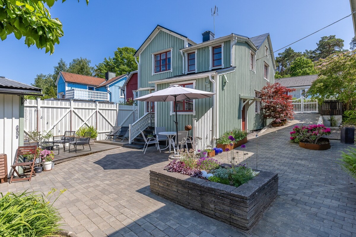 A well-maintained courtyard is shown, featuring a mix of outdoor seating arrangements. A large umbrella provides shade over a dining area set against a backdrop of flowering plants and greenery. The two-story building with wooden siding is visible, complementing the serene atmosphere.