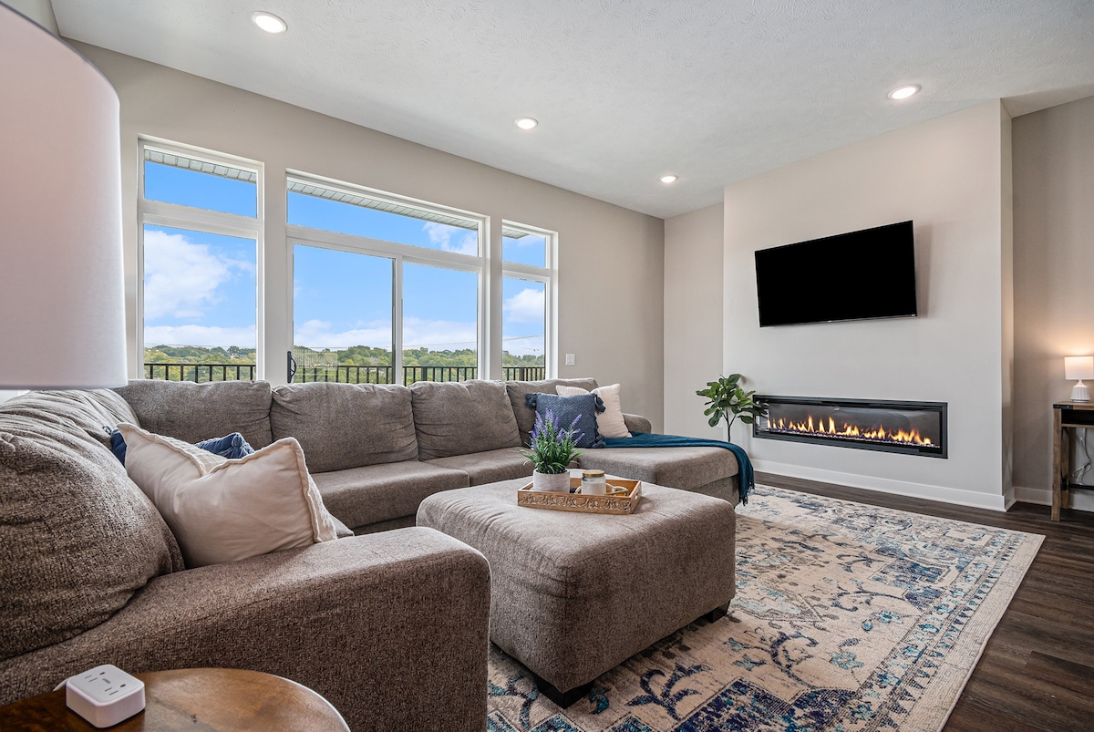 A spacious living area is highlighted by large windows offering scenic city views. A modern gas fireplace is mounted on the wall beneath a flat-screen television. A plush sectional sofa and a decorative area rug create an inviting atmosphere, complemented by natural light streaming in.