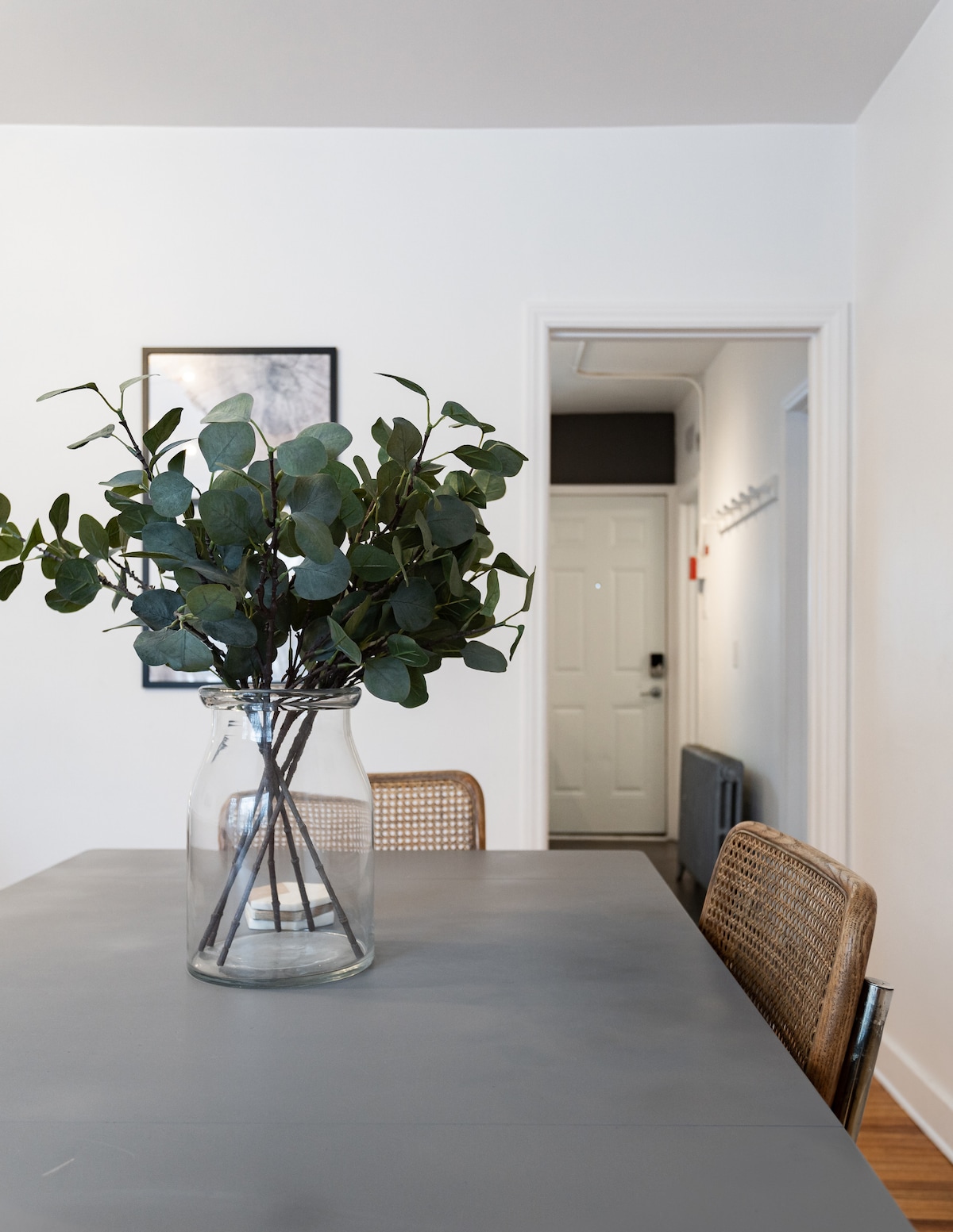 A dining table is showcased in the foreground, featuring a glass vase filled with green foliage. Two textured chairs are positioned at the table. In the background, a door with a simple design leads to another room, enhancing the open space.