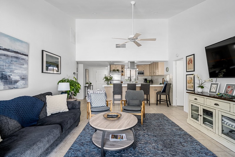 A spacious living area features a dark blue area rug beneath a round coffee table. Cozy seating includes a dark sofa and two armchairs. A ceiling fan and wall-mounted TV enhance comfort, while the open layout allows views into the kitchen area in the background.