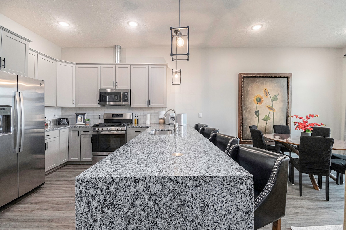 A modern kitchen is showcased with a spacious granite island and stainless steel appliances. The open layout connects to a dining area featuring a wooden table and black leather chairs. Bright overhead lighting enhances the inviting space filled with natural light.