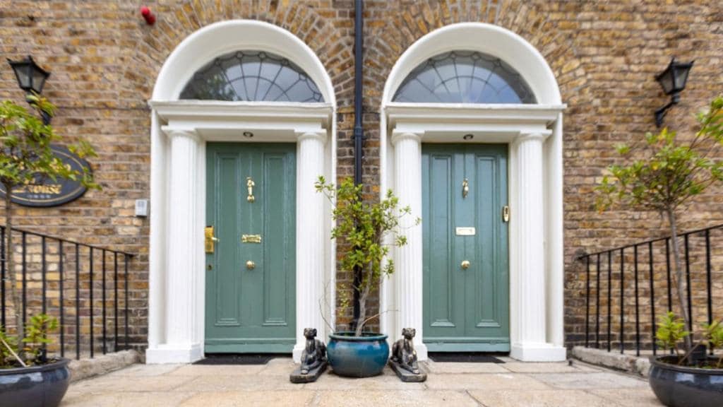 Two elegantly designed entrance doors are framed by white columns, each door painted in a soothing green shade. Flanking the doors are planters with small trees, placed on a stone pathway. Vintage-style wall lamps add a touch of charm to the entrance.
