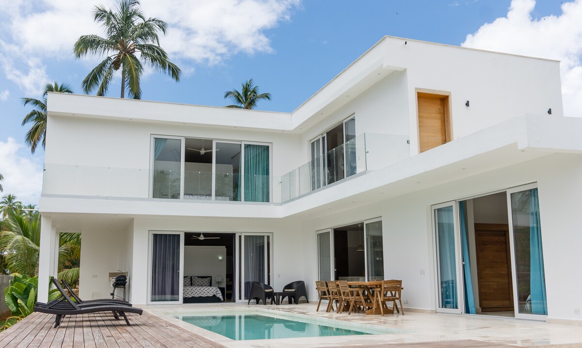 A modern two-story villa is displayed, featuring expansive glass doors that open to the pool area. A private pool is surrounded by a wooden deck with lounge chairs and an outdoor dining set. Lush greenery and palm trees provide natural privacy.