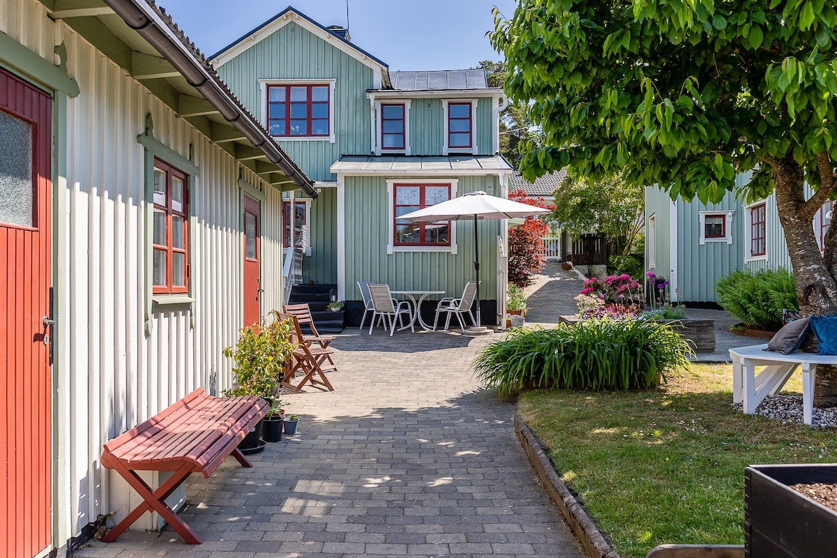 A serene outdoor area is shown, featuring a pathway lined with greenery and blooming flowers. The entrance of the building is flanked by red doors, while a seating arrangement with chairs and an umbrella provides a shaded spot for relaxation.